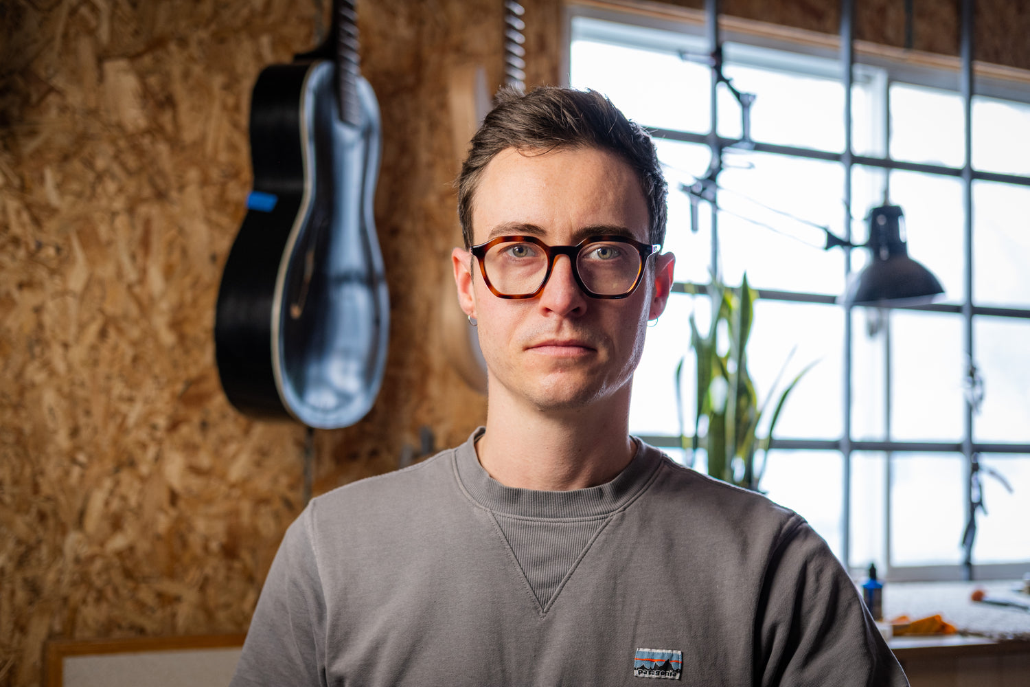 Man wearing glasses and a gray shirt in a room with a guitar and window in the background